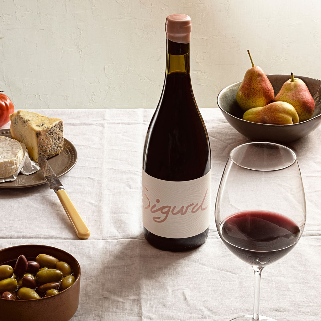 Bottle of Sigurd wines Cabernet with a glass of red wine on a table with cheese, pears, and olives.